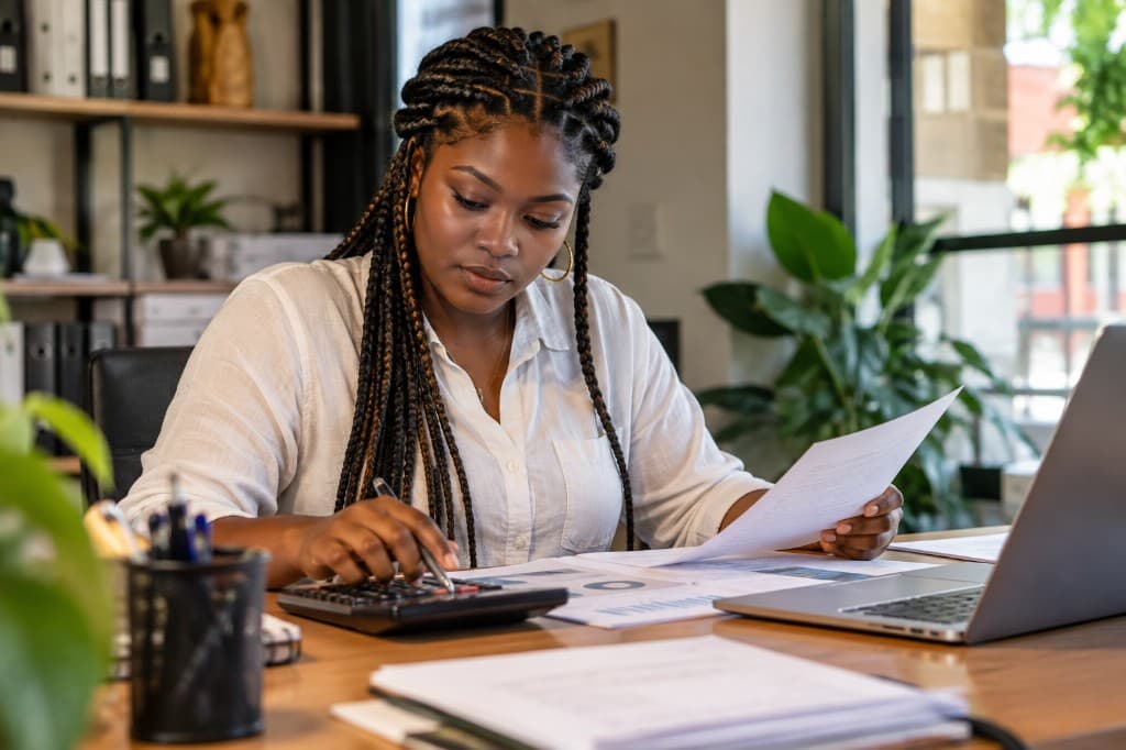 Mujer afrocolombiana con trenzas revisa documentos y hace cuentas en su escritorio de trabajo.